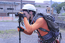 測量機器を覗き込む山本さん（事務所前の駐車場にて）。服装は、林内でも目立つ原色系のもので、スポーツ用もしくは業務用の速乾服を着用している。